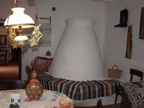A képen a füzesgyarmati tájház egyik helyisége látható búbos kemencével, a falon használati tárgyak, előtérben egy asztal, rajta kulacs, fölötte díszes lámpatest.