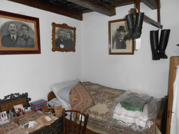 A képen a füzesgyarmati tájház egyik szobája látható vetett ággyal, a falon családi fényképekkel.