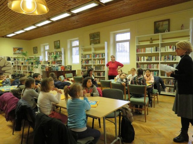 Könyvtári szakkör Egy könyvtárban több asztalnál gyerekek ülnek, a könyvtáros hölgy beszél, háttérben a pedagógus.
